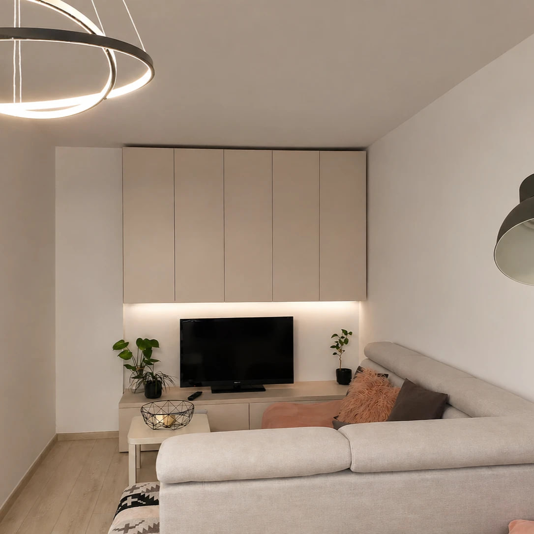 Living room of a small apartment: a pale sectional sofa with blush cushions, a run of cream floor-to-ceiling cabinetry above a low media unit lit from beneath, a ring pendant overhead and a dark angled floor lamp at the right.