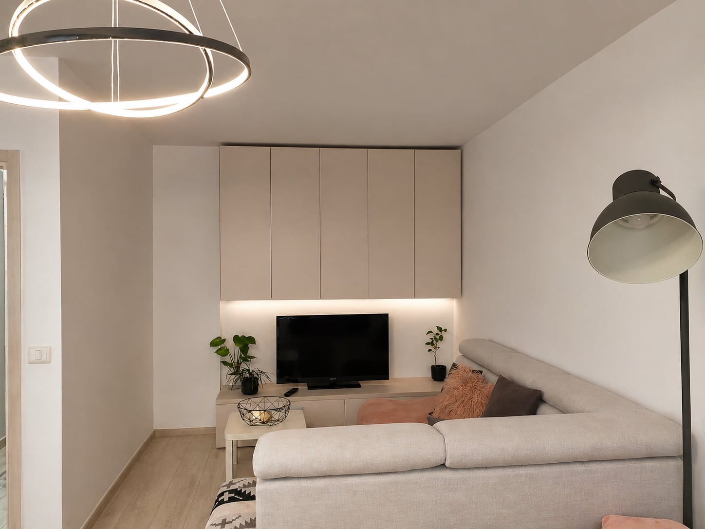 The finished living room: a pale sectional sofa with blush cushions, cream floor-to-ceiling cabinetry above a low media unit lit from beneath, a ring pendant overhead and a dark angled floor lamp.