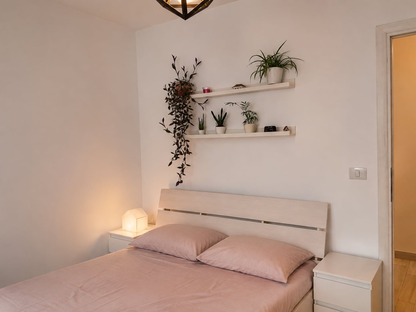 The finished bedroom: two pale shelves of plants above the headboard, a trailing vine, a small house-shaped lamp lit on the bedside table.
