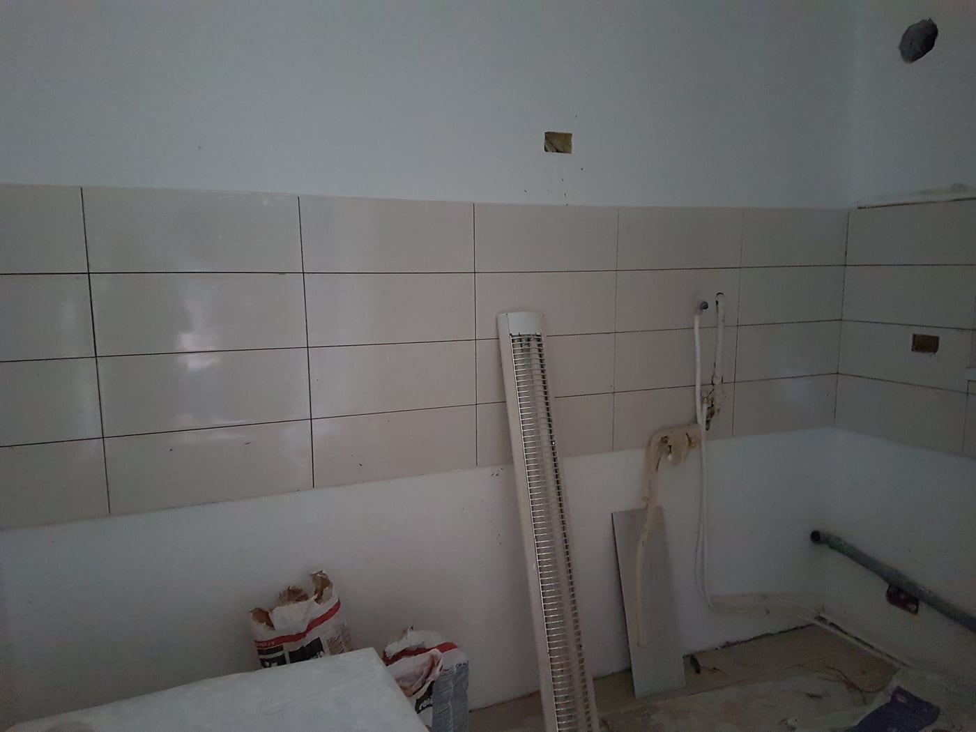 The bathroom mid-works: half-tiled walls, an exposed shower feed, bags of adhesive and a light fitting propped against the bath.