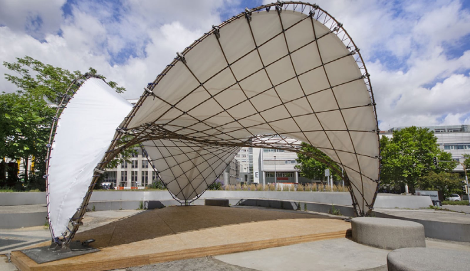 The finished pavilion on the university campus: two curved membrane shells stretched over a lattice of natural fibre profiles, above a timber deck.
